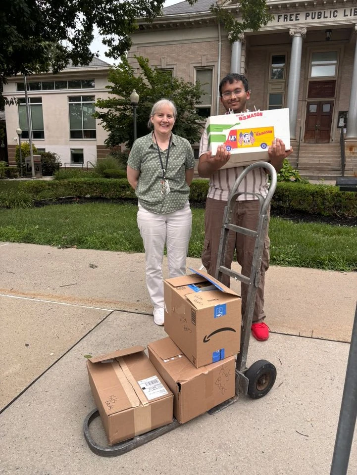 A Pages for Change volunteer with a hand truck of book boxes next to a librarian outside the New Brunswick Free Public Library